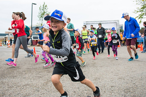 A child in a baseball cap smiles for the camera while running at Carbone's Race for Research, with other runners in the background.