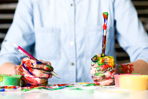 A person with paint on their hands, holding a paint brush and pencil in either hand.