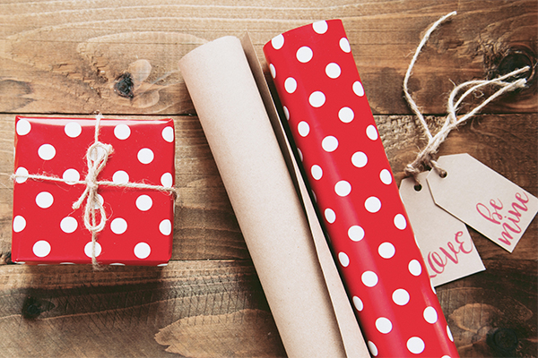 A wrapped gift next to two rolls of wrapping paper and tags reading "Love" and "Be Mine". By Mira Bozhko.