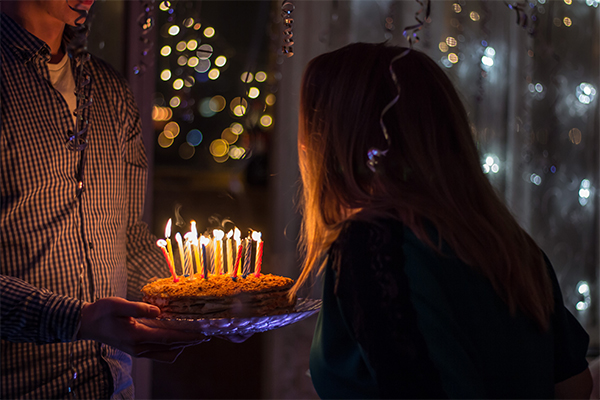 One person holding a cake with lit candles on it so that a second person can blow out the candles. By Sergei Solovev.